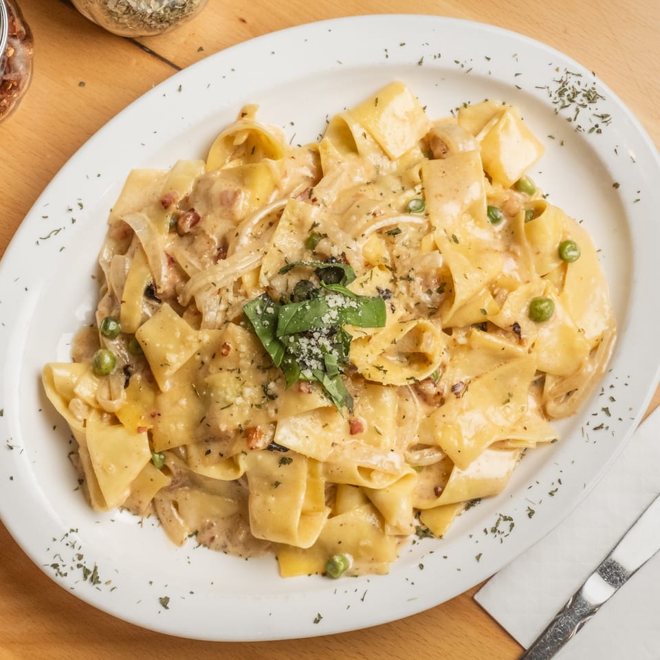 Creamy ribbon pasta with peas, bacon, herbs, basil, and grated cheese served on a white plate.