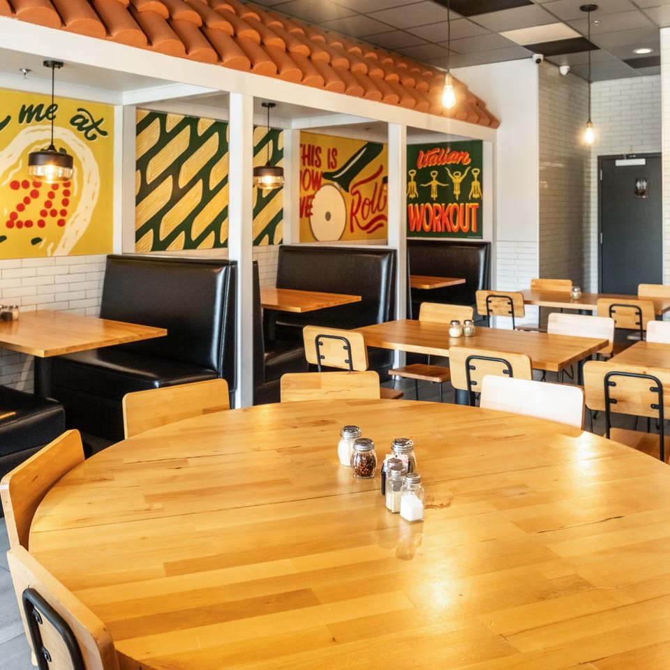 Bright Italian restaurant dining room with wooden tables, black booths, pendant lights, colorful murals, and terracotta roof decor.
