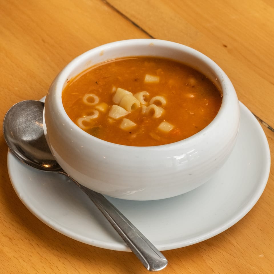 Bowl of tomato soup with ring-shaped pasta and diced vegetables on a white saucer, accompanied by a spoon on a wooden table.