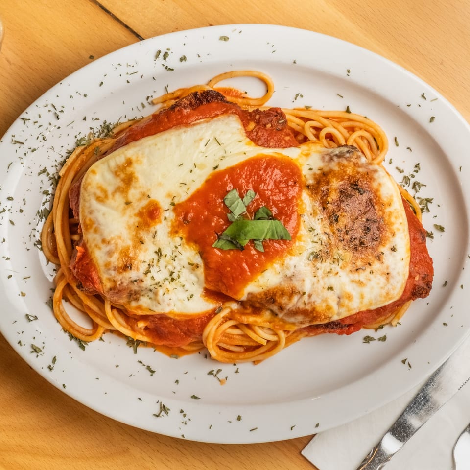 Baked chicken Parmesan with melted mozzarella, marinara sauce, and basil served over spaghetti on a white plate.