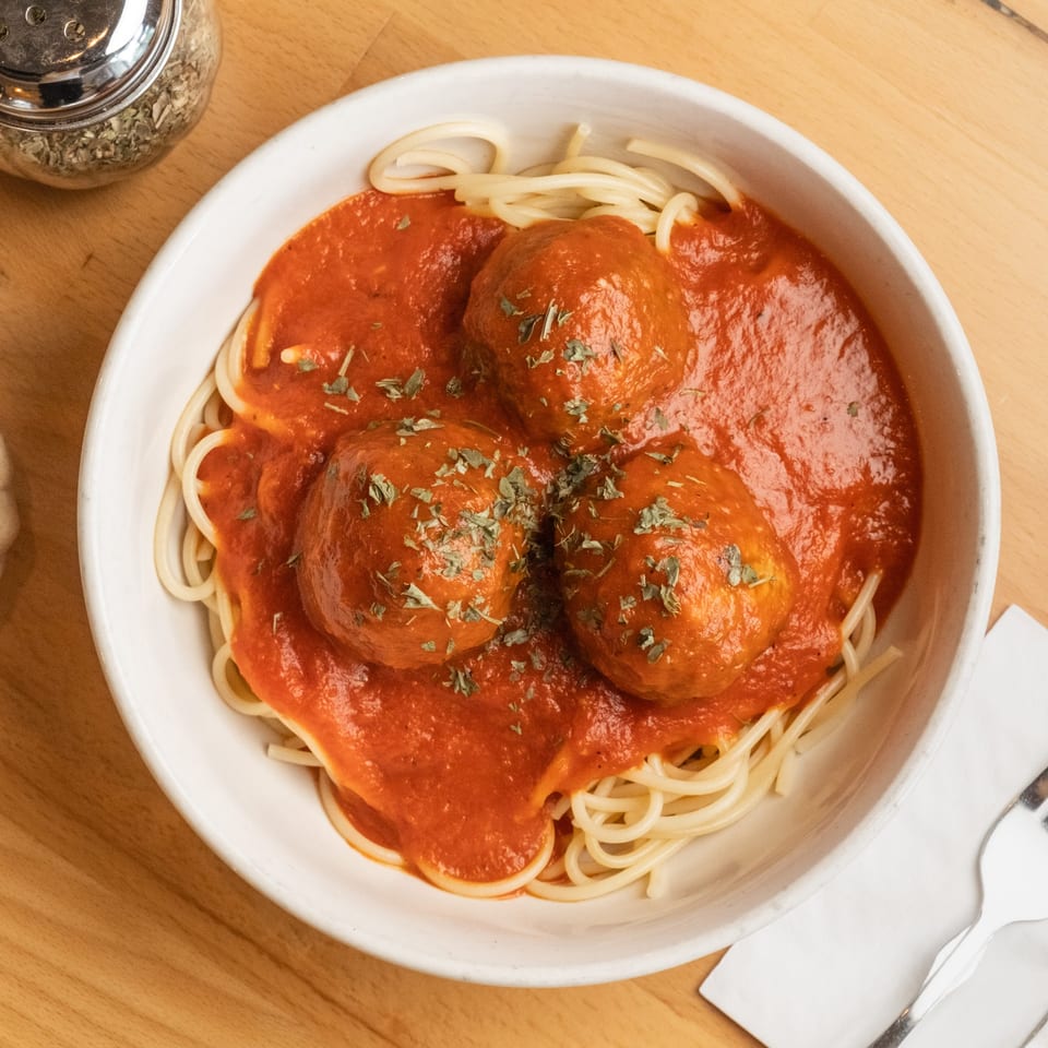 Bowl of spaghetti topped with four meatballs, tomato sauce, and herbs on a wooden restaurant table.