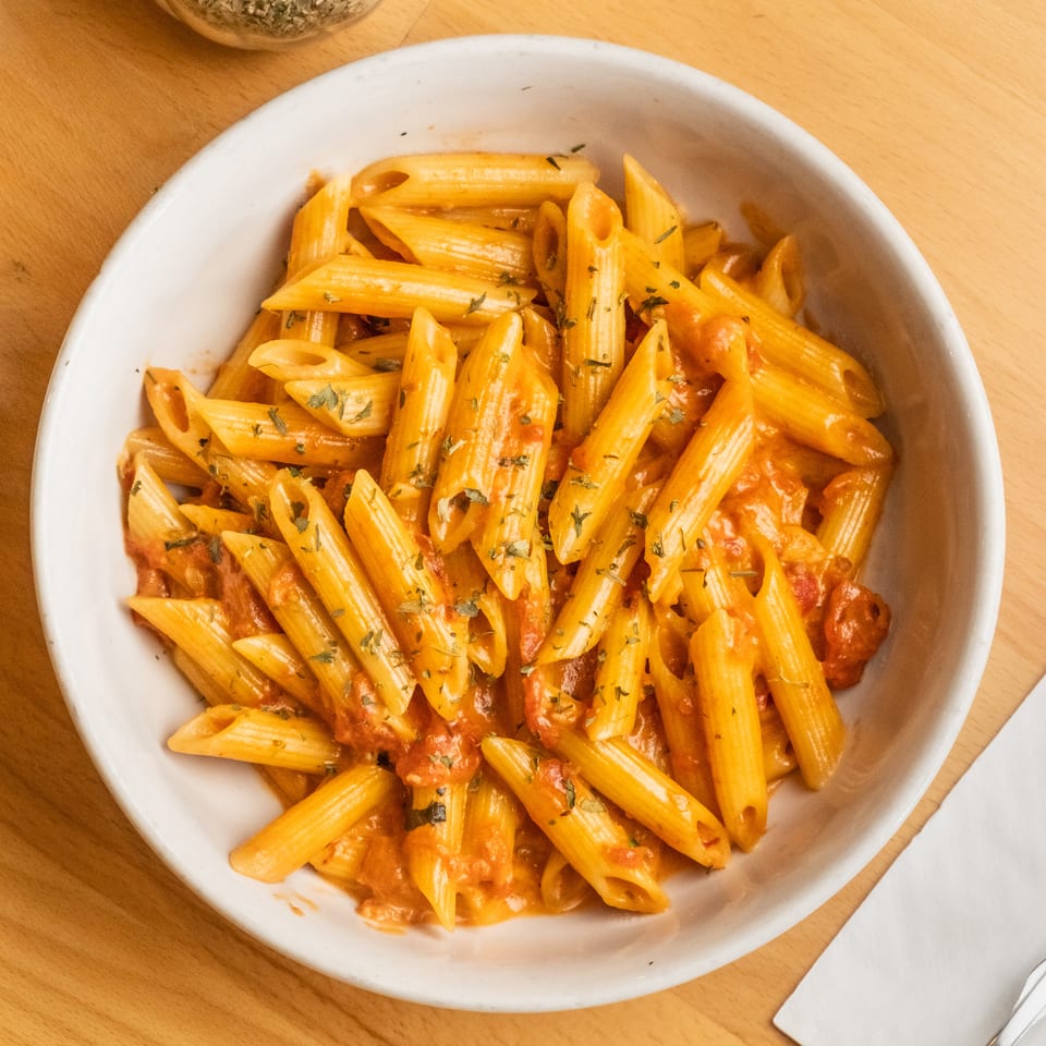 Bowl of penne pasta in tomato sauce, garnished with herbs, on a wooden restaurant table.
