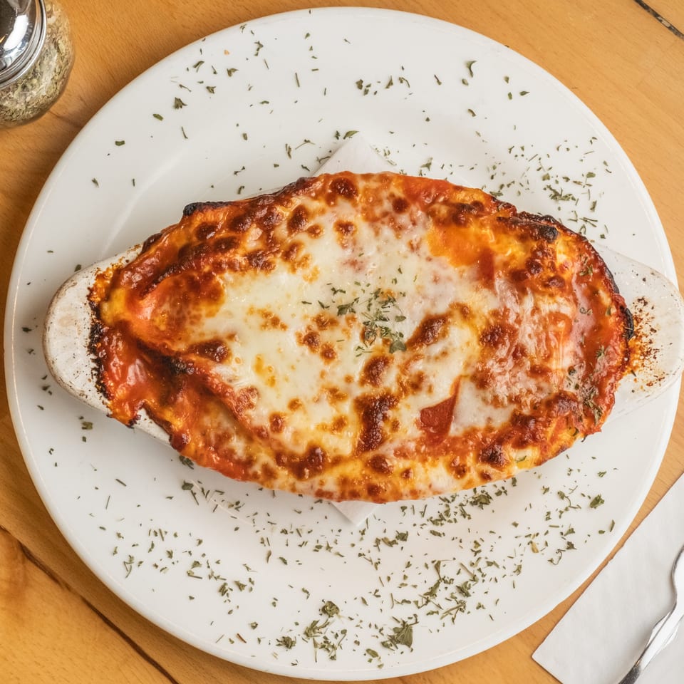 Baked pasta dish covered with browned melted cheese and herbs, served on a white plate atop a wooden restaurant table.