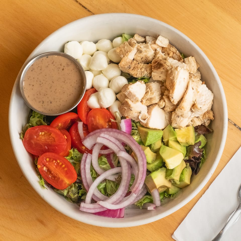 Overhead view of a salad topped with grilled chicken, tomatoes, avocado, red onion, mozzarella, and creamy dressing.