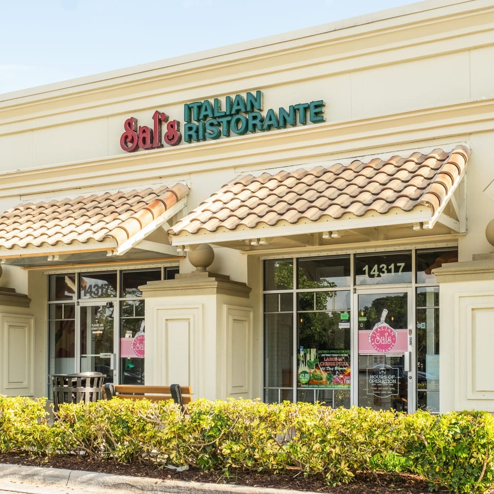Exterior of Sal’s Italian Ristorante featuring cream stucco, terracotta tile awnings, glass doors, and landscaped hedges.