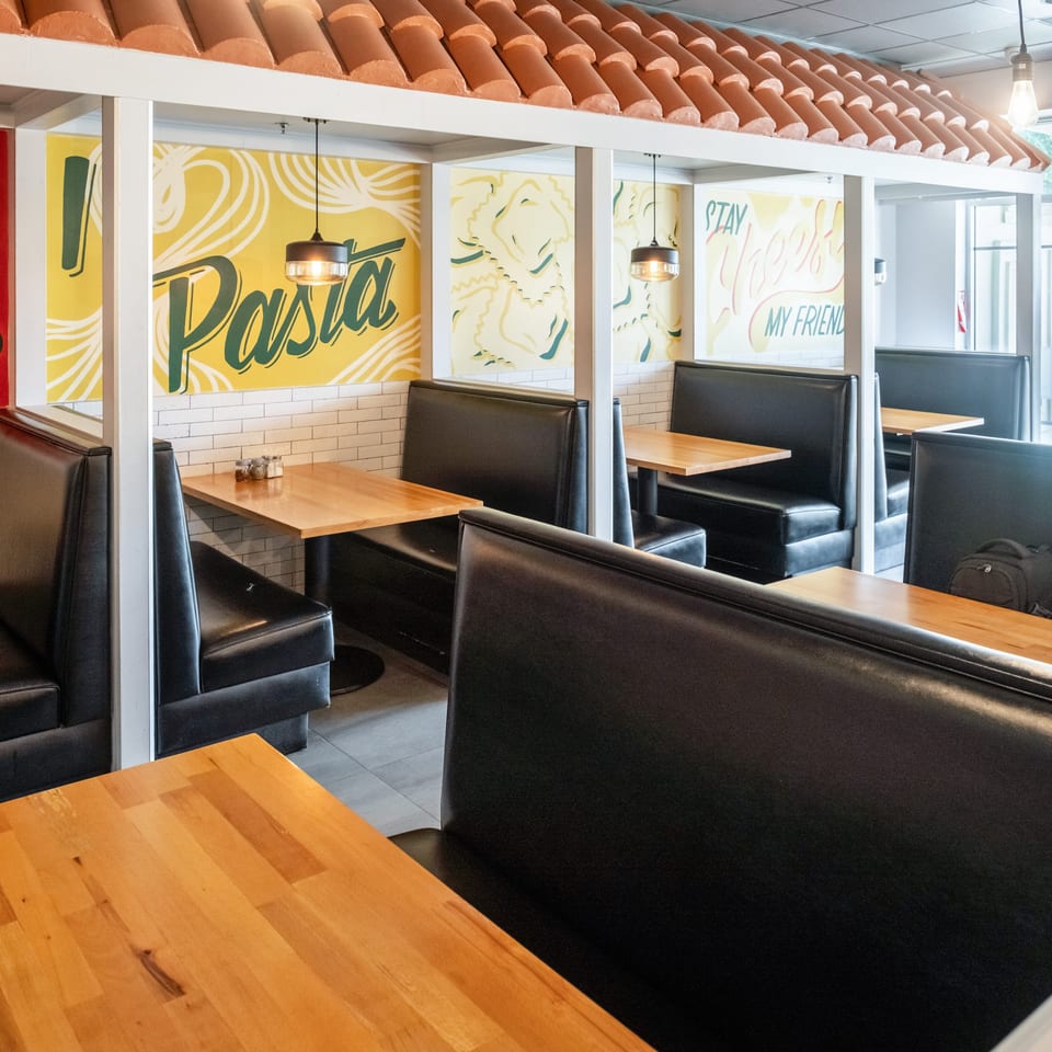 Bright restaurant dining area with black leather booths, wooden tables, pendant lights, colorful murals, and a tiled Spanish-style ceiling.